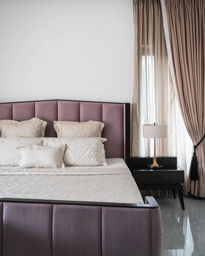 Mauve upholstered bed with cream pillows beside sheer curtains and a dark wood nightstand