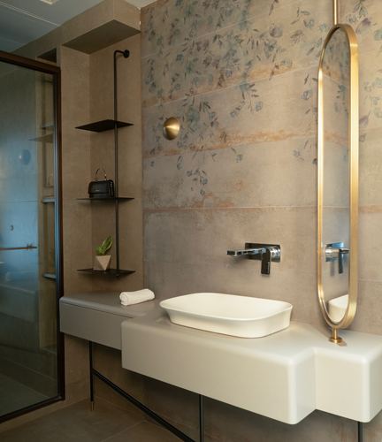 Bathroom with floral-patterned stone wall, sculptural white vanity, and oval brass mirror