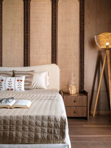 Bed with cane-panelled headboard beside wood-clad wall and tripod floor lamp with wire cage shade