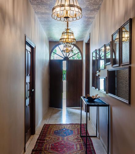Narrow hallway with hanging chandeliers, arched wooden door, mirrors, and a patterned runner rug
