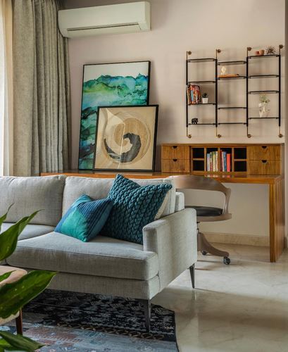 Living room corner with grey sofa, teal cushions, leaning artwork, and wooden desk with black wall shelf