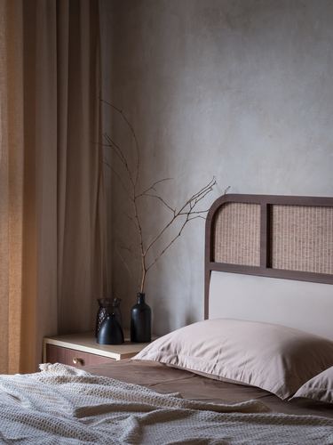 Bedroom corner with cane headboard, linen curtains, and dried branch in black vase on nightstand