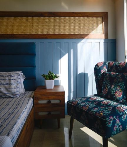Sunlit bedroom corner with cane headboard, blue panelled wall, wooden nightstand and floral armchair