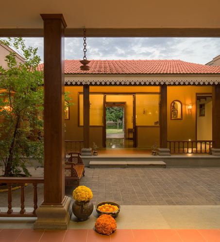 Twilight view through teak columns of a traditional courtyard house with sloping tile roof and marigold offerings