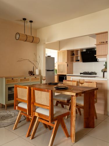 Wooden dining table with cane-back chairs beneath cylindrical cane pendant lights, kitchen visible beyond