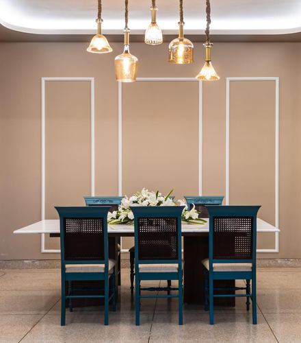 Dining table with teal cane-back chairs under cluster of amber pendant lights against beige paneled wall