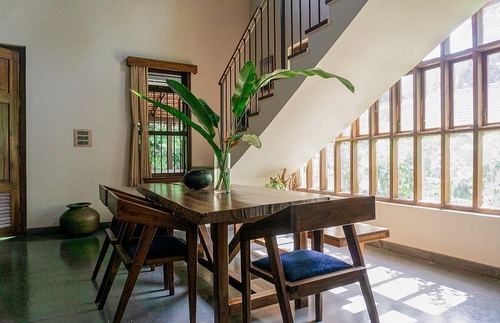 Wooden dining table with blue-cushioned chairs beneath a staircase, large grid windows lighting the room
