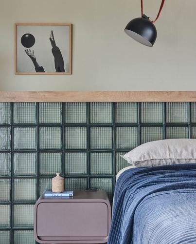 Bedroom corner with glass block wall, wood trim, black pendant lamp and mauve nightstand beside a blue bed