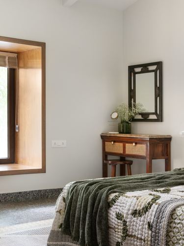 Bedroom corner with cane-front console, antique mirror, and green quilted bedspread near a wooden window nook