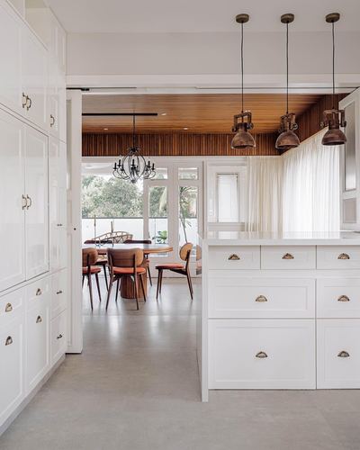 White kitchen island with brass pendant lights opening to a wood-ceilinged dining area