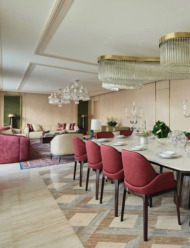Open-plan dining and living room with burgundy velvet chairs, brass chandeliers, and marble floor.