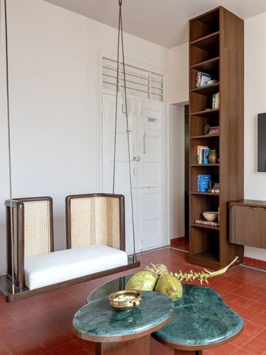 Cane swing bench beside tall wood bookshelf, terracotta floor, oval stone coffee table with coconuts