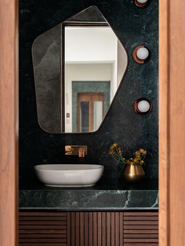 Powder room vanity with dark green stone wall, irregular mirror, and brass fittings framed by wood door