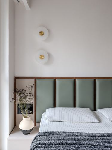 Bedroom with sage leather-paneled headboard, twin round wall sconces, and ceramic vase with dried flowers