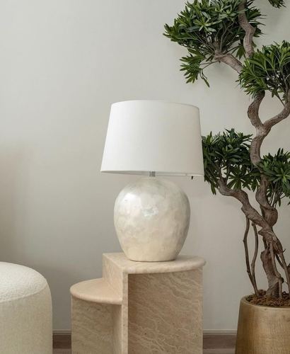 Cream ceramic table lamp on a two-tiered travertine side table beside a boucle ottoman and potted tree