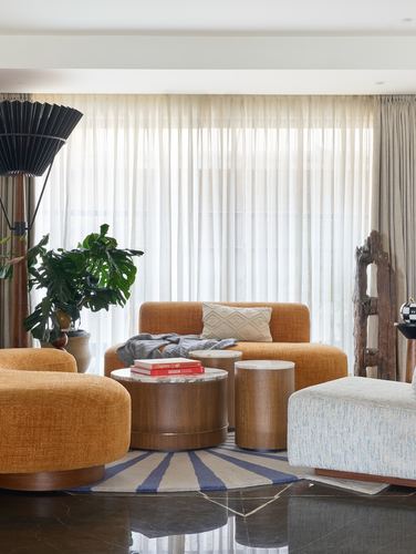 Curved orange sofas around round wood and marble tables on a striped rug before sheer curtains