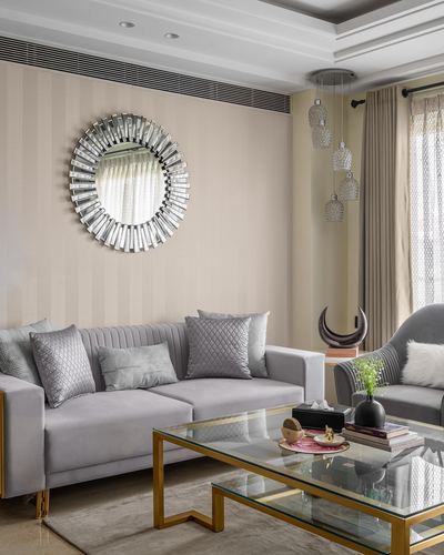 Grey velvet sofa beneath a sunburst mirror in a pastel neutral living room with gold-framed glass table
