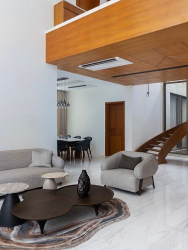 Double-height living room with wood-clad overhang, curved staircase, and grey sofas on marble floor
