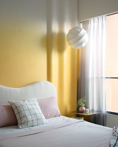 Bedroom corner with mustard wall, curved white headboard, pink bedding, and round pendant light