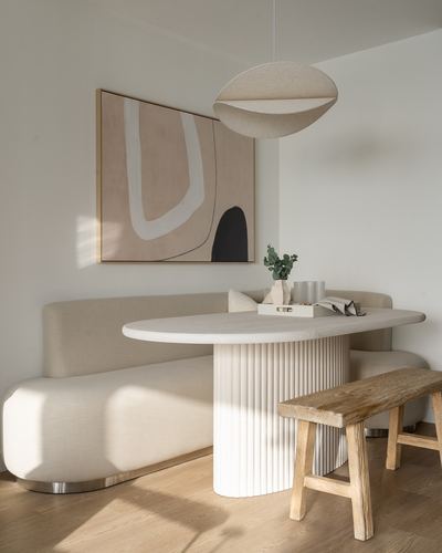 Curved beige banquette and fluted stone dining table beneath abstract art and sculptural pendant light