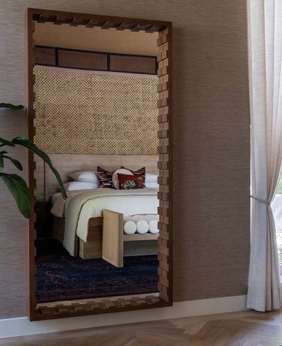 Bedroom reflected in a large carved wooden mirror beside a linen curtain and plant