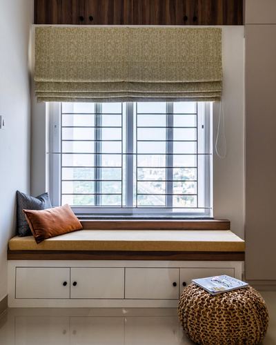 Window seat with mustard cushion, patterned blind, and knit pouf beside grilled window overlooking city