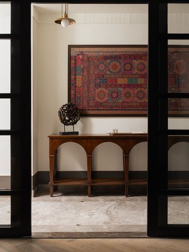View through black-framed glass doors into a foyer with an arched wooden console and patterned wall art
