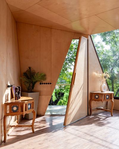 Angular plywood-lined room with triangular window opening to greenery and carved wooden console tables