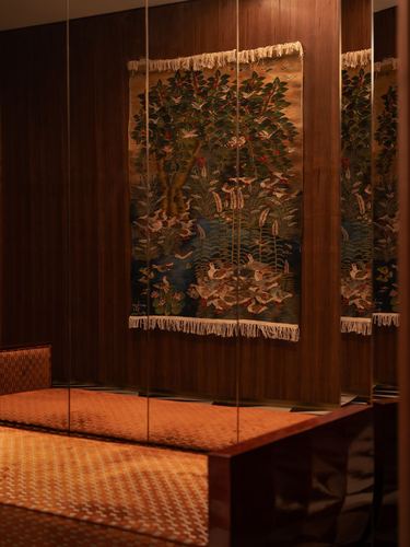 Wood-paneled bedroom corner with a bird-and-tree tapestry and woven orange bedspread reflected in mirrored panels