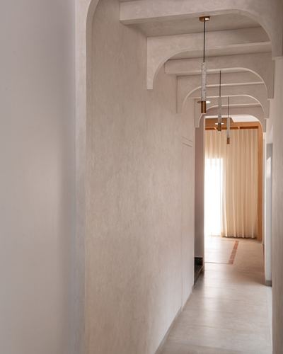 Curved plastered corridor with arched beams, hanging pendant lights, leading to a sunlit curtained doorway.