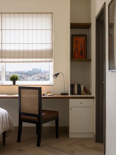 Study nook with wooden desk, cane-back chair, window with roman blind, and built-in shelving with art