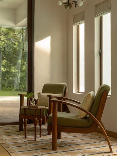 Two curved teak armchairs with olive cushions beside tall windows and a garden view