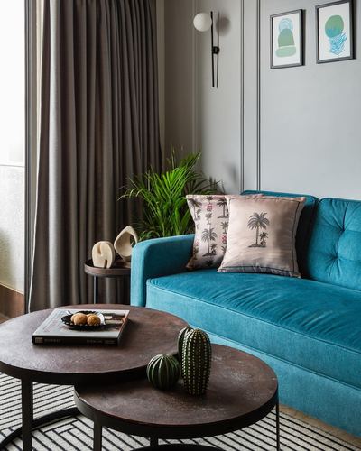 Teal velvet sofa with palm-print cushions beside round metal coffee tables and paneled grey wall