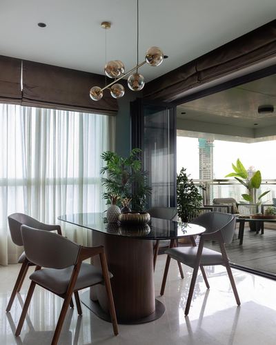 Oval glass-top dining table with grey chairs under a brass chandelier, balcony view beyond