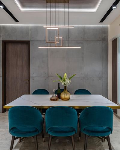 Dining table with teal velvet chairs beneath a geometric pendant light against a concrete panel wall