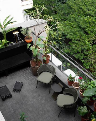 Elevated view of a grey-tiled balcony with woven olive chairs, potted plants and a small water feature, framed by trees.