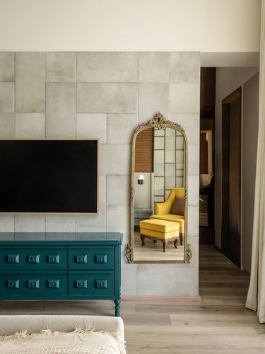 Teal cabinet below wall tv, gilded arched mirror reflecting a yellow armchair, textured stone wall