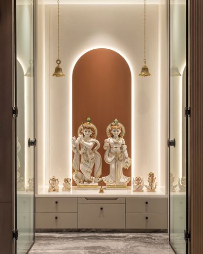 Pooja niche with arched terracotta backdrop, glowing cove lighting, and marble deity statues on a cabinet