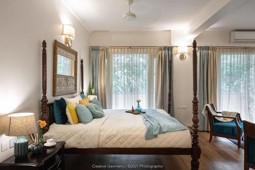 Bedroom with carved wooden four-poster bed, teal and mustard cushions, sheer curtained windows and wood flooring