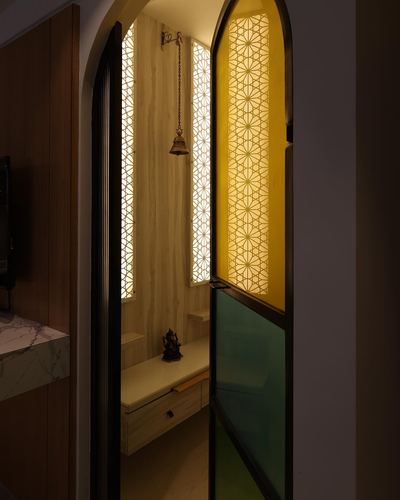Arched pooja room doorway with backlit jali panels, brass bell and colored glass door