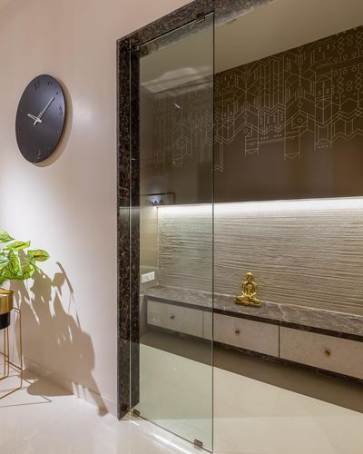 Pooja niche with brass buddha idol behind glass door, textured stone wall, marble clock on foyer wall
