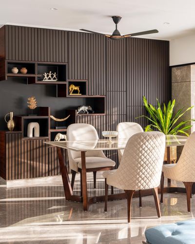 Dining area with fluted dark wood wall, cubbyhole shelving with curios, and quilted cream chairs around a stone table