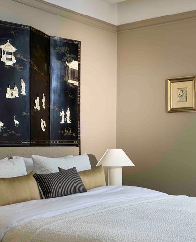 Bedroom corner with black lacquer inlaid screen behind bed, white lamp, framed art on beige wall