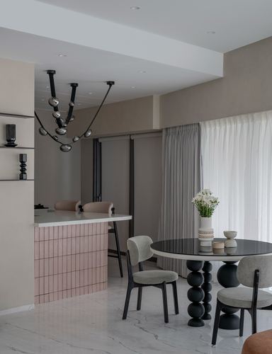 Round black dining table with grey chairs beside a pink-tiled kitchen counter under a sculptural pendant light