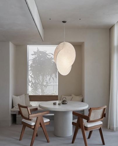 Round stone dining table with cane armchairs beneath a large sculptural pendant light in a niche