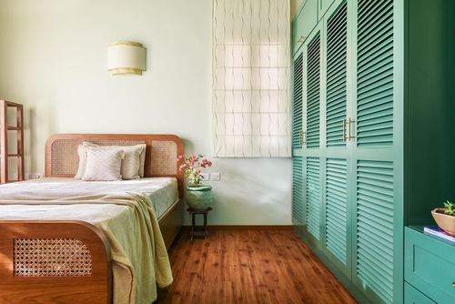 Bedroom with cane headboard bed, wood floor, and teal louvered wardrobe by a curtained window