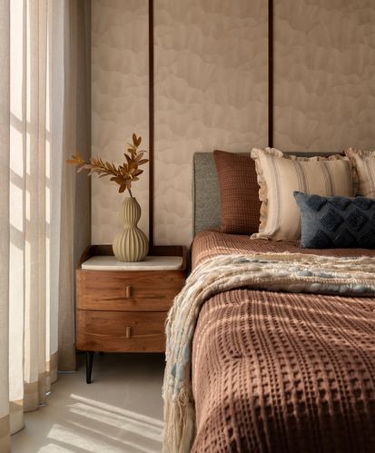 Sunlit bedroom corner with walnut nightstand, gourd vase, and layered terracotta bedding against panelled wall