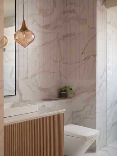 Marble-clad bathroom corner with amber glass pendant, fluted wood vanity, and floating shelf