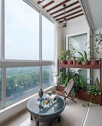 Balcony sit-out with round glass table, folding chair, potted plants, and floor-to-ceiling windows overlooking greenery