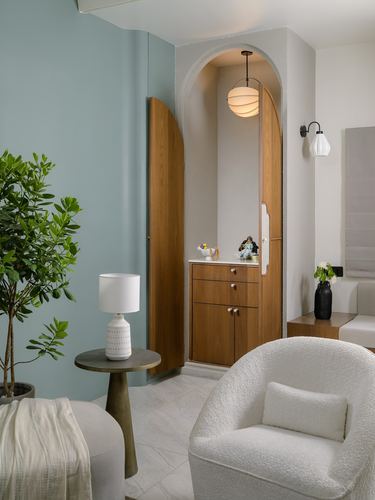 Arched wooden cabinet niche with pooja idols beside a sage green wall and boucle armchair